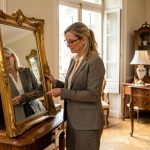 Une antiquaire examine un miroir doré dans un élégant showroom parisien entouré de meubles anciens et d’objets de valeur.